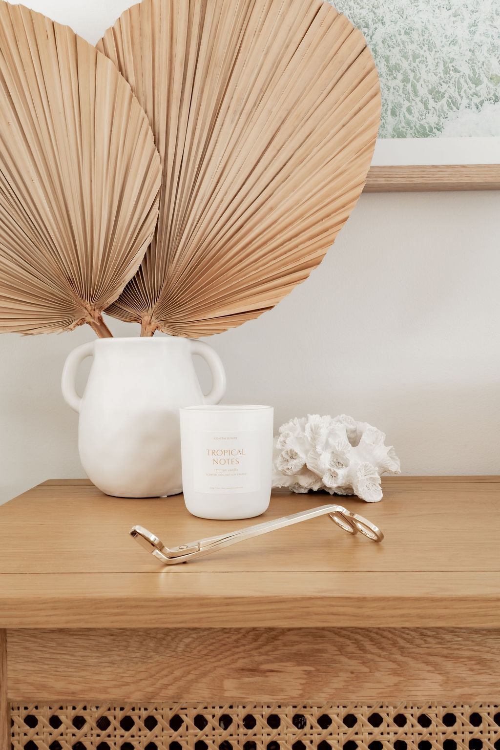Tropical Notes white candle on a wooden surface, accompanied by a white vase and dried palm leaves.